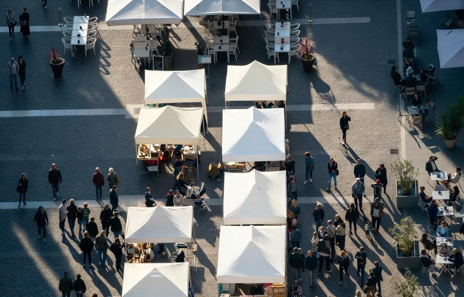 Vogelperspektive auf einen belebten Marktplatz mit einer Reihe von weißen und beigen Faltpavillons auf Kopfsteinpflaster. Menschen flanieren bei hellem Tageslicht zwischen den Marktständen, die lange Schatten werfen.